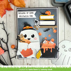 A Halloween-themed layered card or craft design displayed on a light wood surface with autumn leaves and spooky props around it. The central artwork shows a smiling white ghost wearing a black witch hat, a black cauldron with orange potion, books, spiderweb, bats, and the text 'BREWING UP SOME HALLOWEEN FUN'.