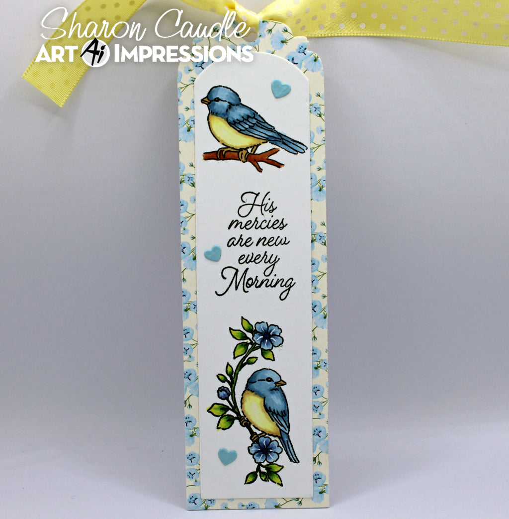 A bookmark with a floral border, featuring two blue-and-yellow birds, small blue hearts, and the phrase “His mercies are new every morning.” A yellow ribbon is tied at the top