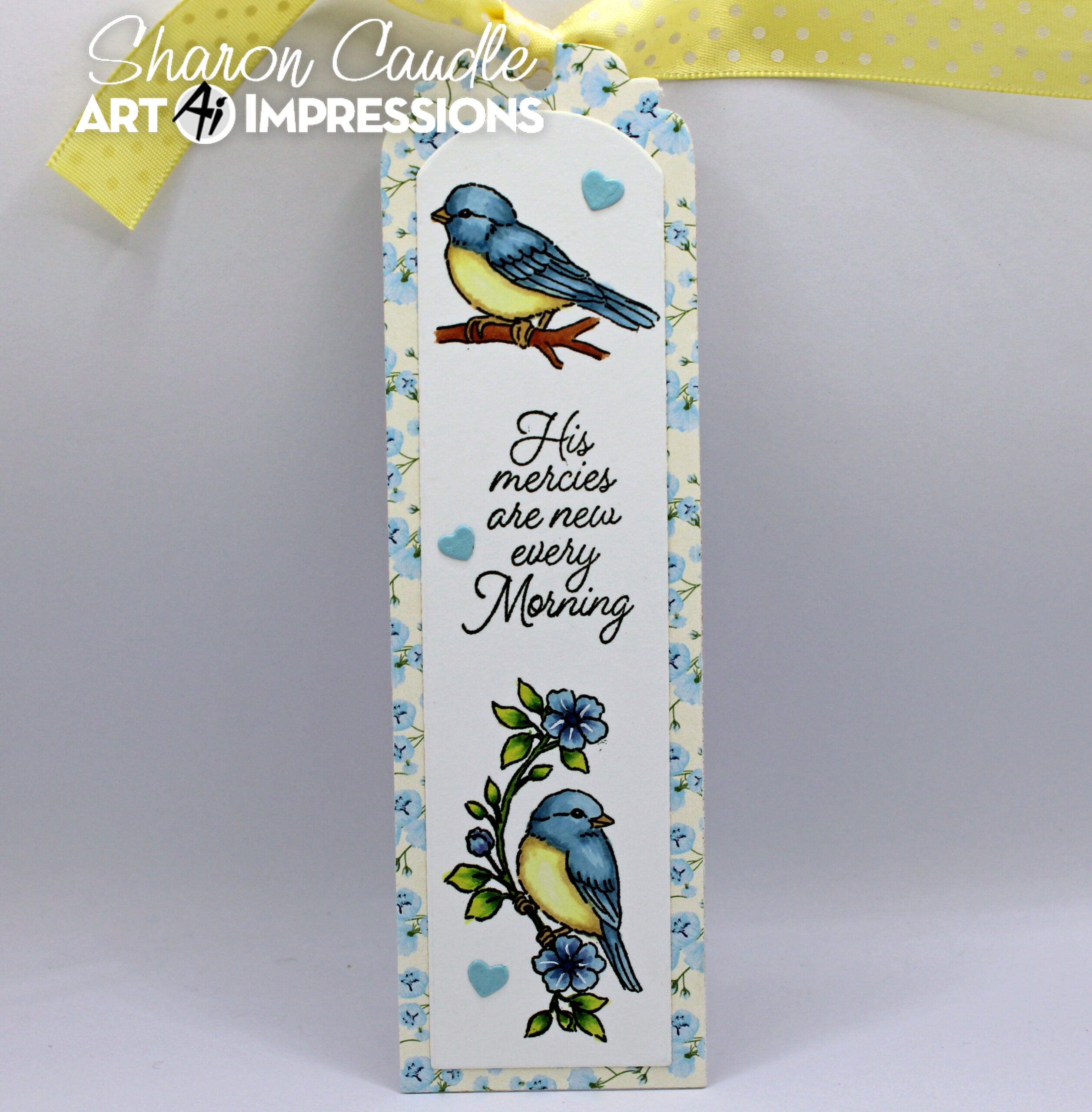 A bookmark with a floral border, featuring two blue-and-yellow birds, small blue hearts, and the phrase “His mercies are new every morning.” A yellow ribbon is tied at the top