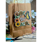 A brown kraft paper gift bag decorated with a floral and butterfly sticker design that reads 'Thank You,' shown with scissors and flowers around it on a tabletop.