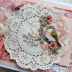 A close-up of a handmade card featuring a cream lace doily with the phrase 'Have Mercy on us God' in brown script, layered over a pale pink background. A die-cut bird illustration with red flowers and green leaves is attached on the right, with a pink ribbon and textured paper accents around it.