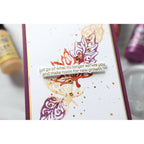 A close-up of a handmade greeting card featuring a white background with purple and orange abstract leaf designs, small gold glitter dots, and a central white banner with the quote, 'Let go of what no longer serves you and make room for new growth.' The card has a deep purple border and is photographed with paint tubes blurred in the background. | color-code:ALT01