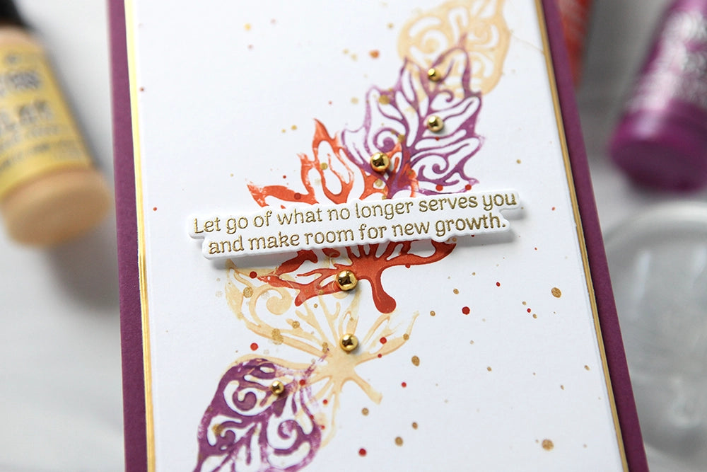 A close-up of a handmade greeting card featuring a white background with purple and orange abstract leaf designs, small gold glitter dots, and a central white banner with the quote, 'Let go of what no longer serves you and make room for new growth.' The card has a deep purple border and is photographed with paint tubes blurred in the background. | color-code:ALT01