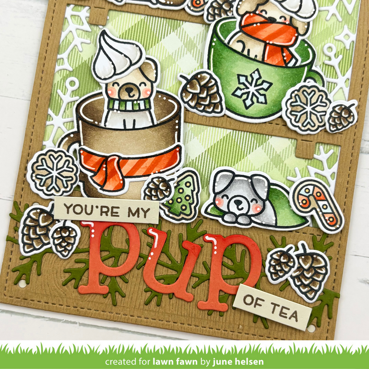 A close-up of a handmade greeting card featuring two cute illustrated dogs sitting in teacups with orange scarves, surrounded by pinecones, snowflakes, and the phrases 'YOU'RE MY' and 'OF TEA'. The card is made from tan cardstock with green patterned paper and red layered lettering.