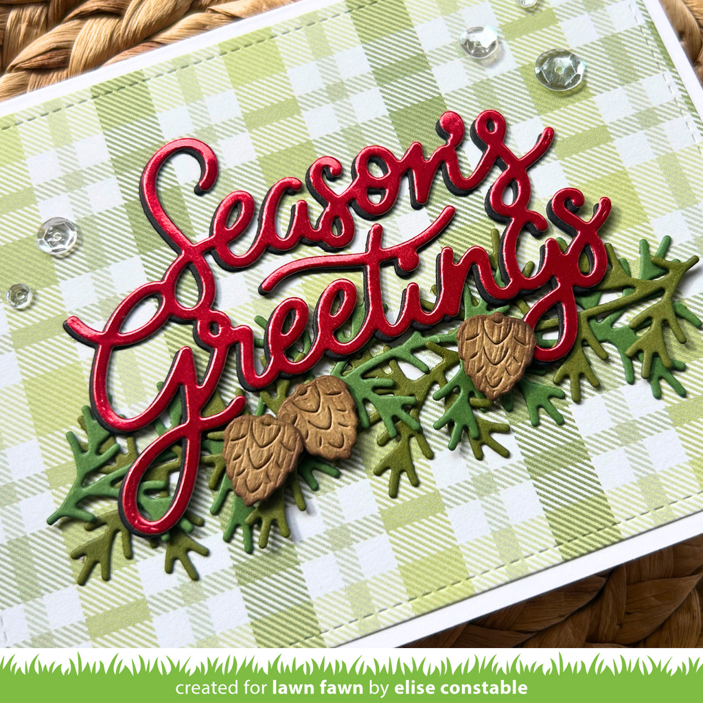 A close-up of a handmade holiday greeting card featuring the words 'Seasons Greetings' in red script over a green and white gingham background, with layered green pine branches and two small pinecones. A green banner at the bottom reads 'created for lawn fawn by elise constable'.