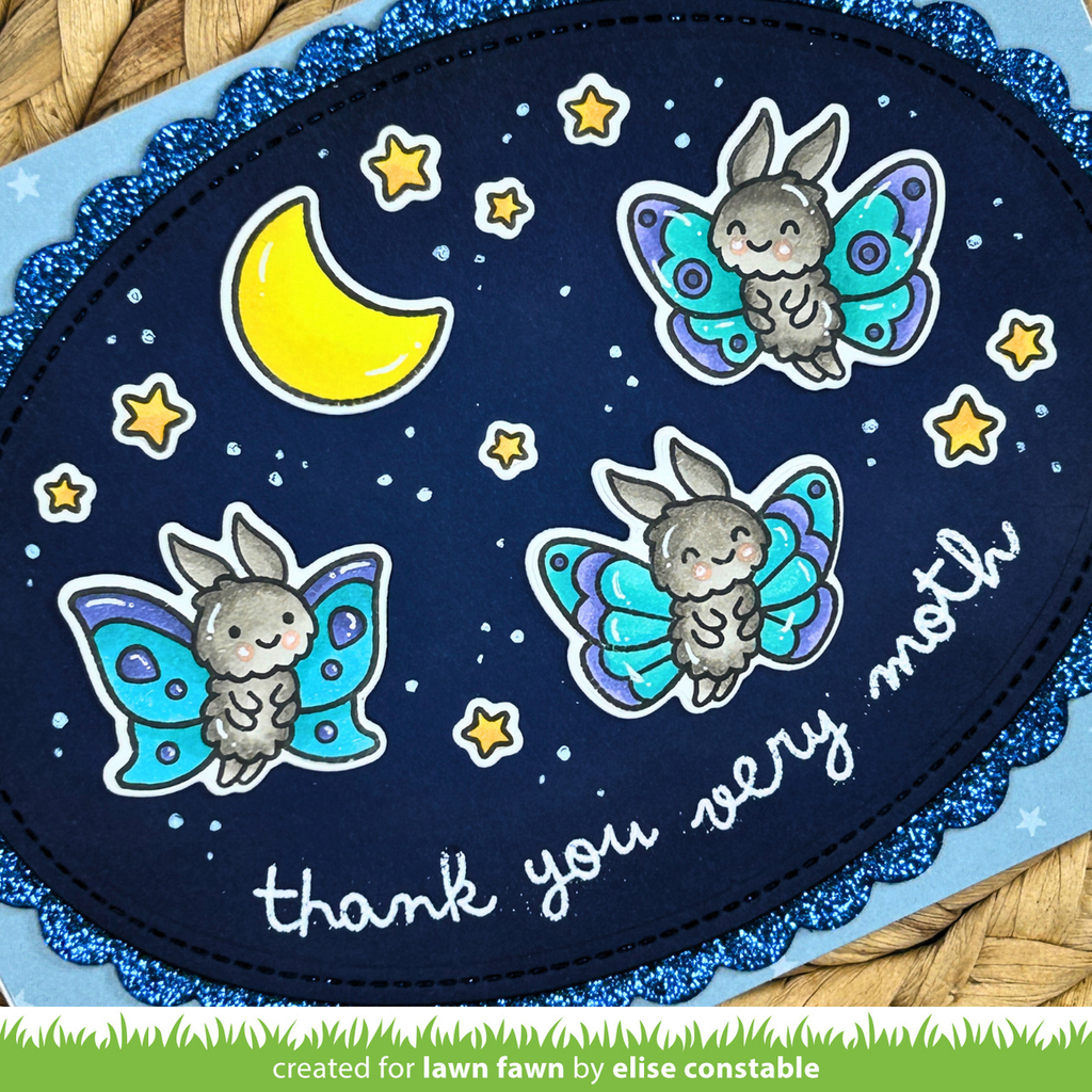 A close-up of a handmade oval card featuring three cartoon rabbits with colorful wings, a yellow crescent moon, stars, and the phrase 'thank you very moth' on a dark blue background with glittery blue scalloped edges. | color-code:alt1