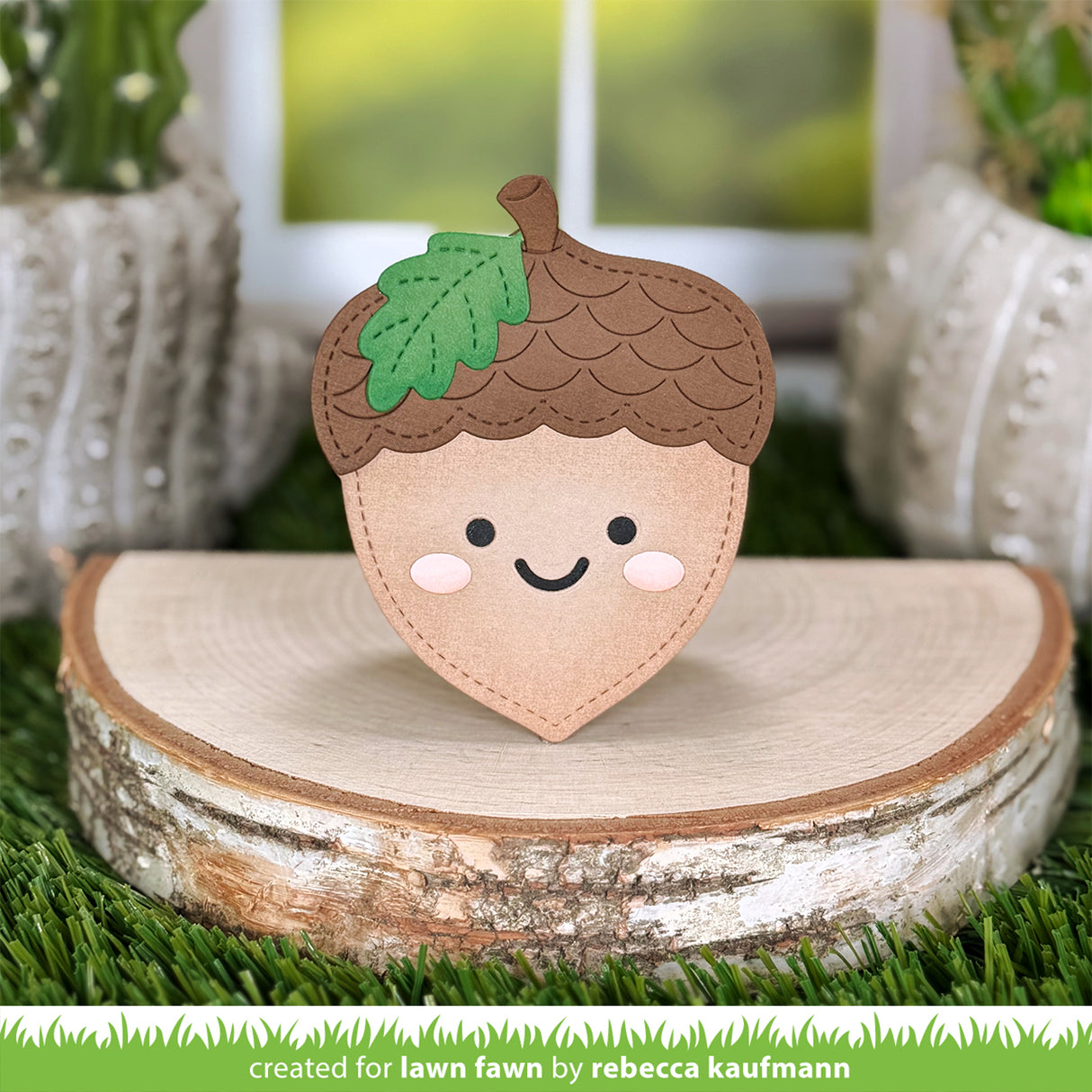A cute, smiling acorn-shaped lawn fawn toy made from felt, displayed on a rustic birch log slice with artificial grass and potted plants in the background.