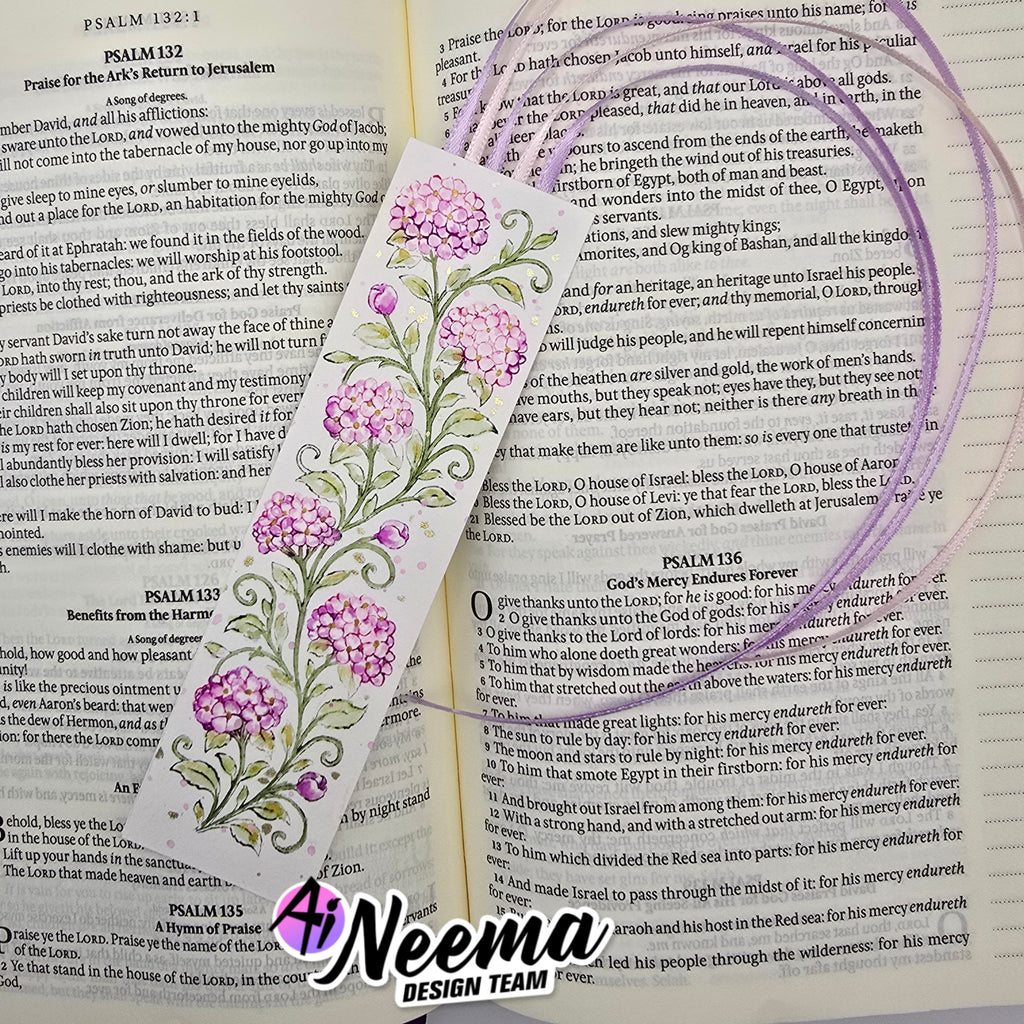 A decorative Bible bookmark featuring a purple and lavender floral vine design with green leaves and white scrollwork, laid over an open book with printed text.