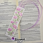 A decorative Bible bookmark featuring a purple and lavender floral vine design with green leaves and white scrollwork, laid over an open book with printed text.