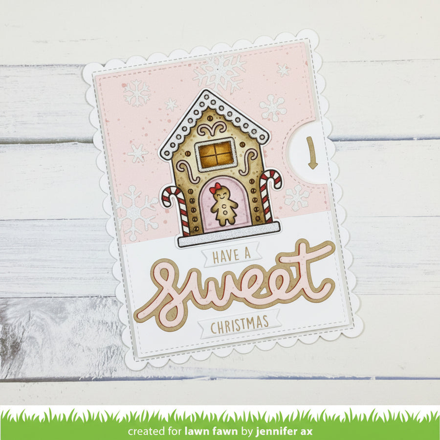 A decorative holiday card with a scalloped white border, featuring a gingerbread house illustration and the phrase “HAVE A Sweet CHRISTMAS.” The card is photographed on a white wood surface.