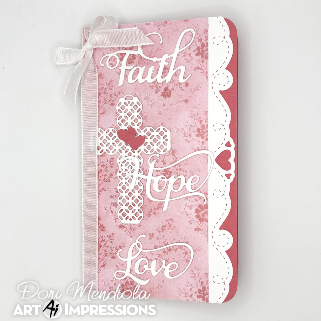 A decorative journal cover featuring the words 'Faith', 'Hope', and 'Love' in white script over a pink floral and damask pattern, with a white lace cross motif and a white ribbon bow at the top, photographed on a light gray background. 