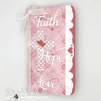 A decorative journal cover featuring the words 'Faith', 'Hope', and 'Love' in white script over a pink floral and damask pattern, with a white lace cross motif and a white ribbon bow at the top, photographed on a light gray background. 