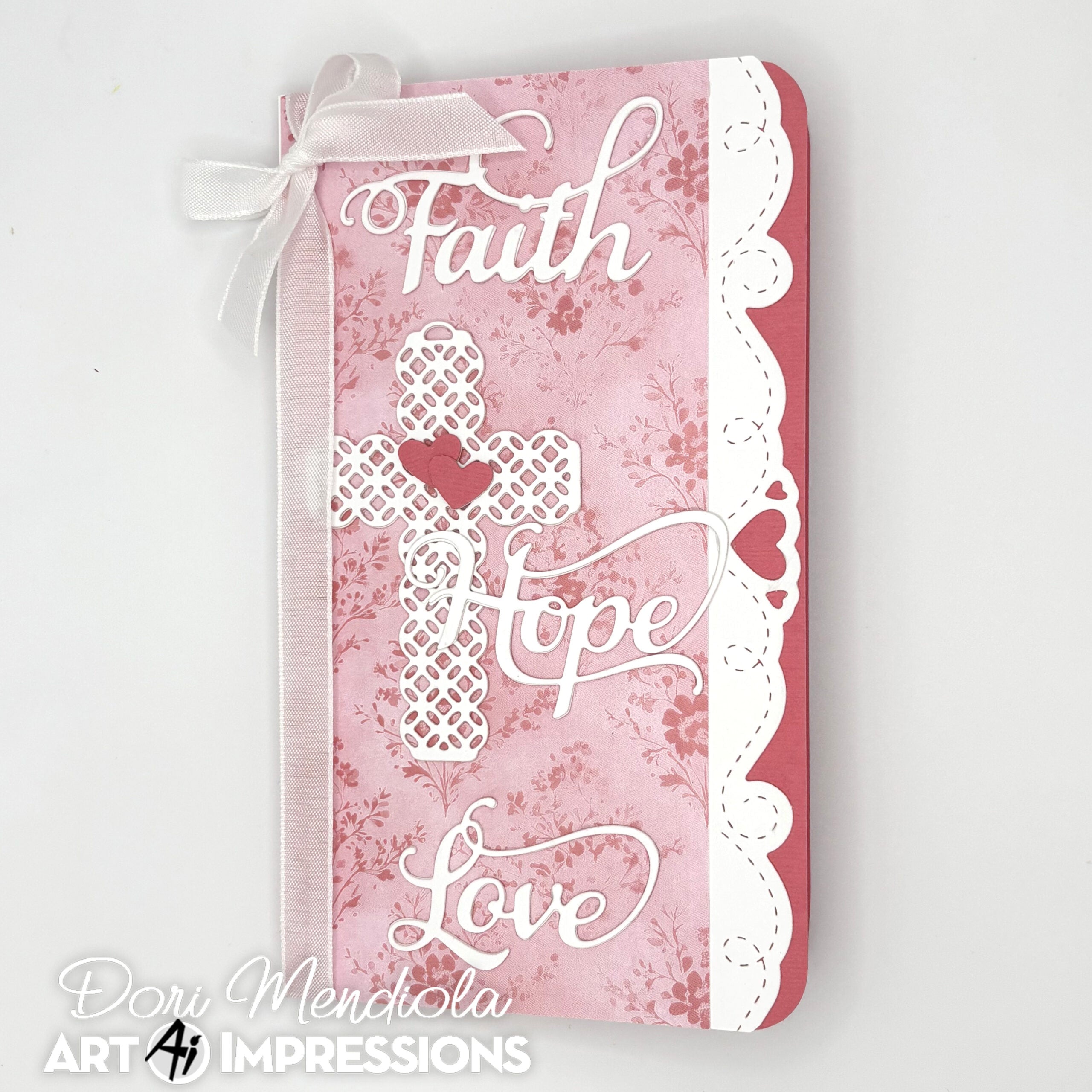 A decorative journal cover featuring the words 'Faith', 'Hope', and 'Love' in white script over a pink floral and damask pattern, with a white lace cross motif and a white ribbon bow at the top, photographed on a light gray background. 
