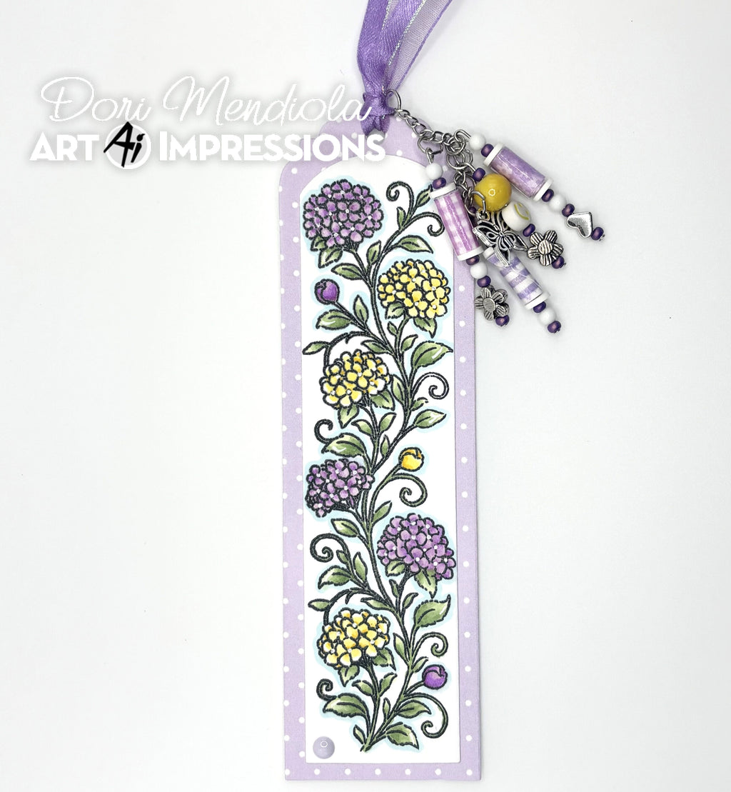 A decorative paper bookmark featuring a hand-drawn purple and yellow floral vine pattern on a white background with a light purple border. The bookmark is adorned with a purple ribbon loop, silver-toned metal charms, and colorful beads