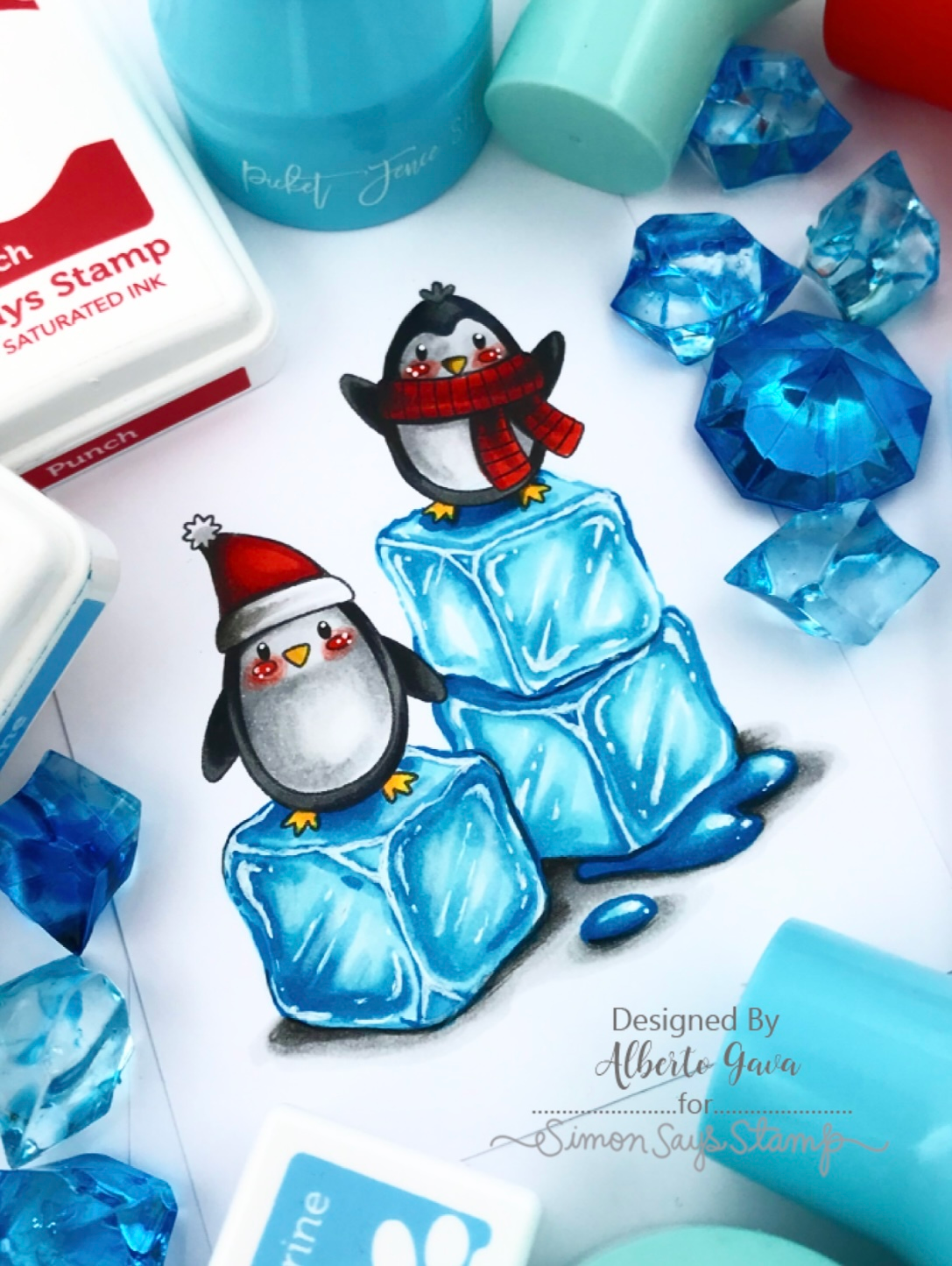 A hand colored design featuring two penguins wearing red Santa hats and scarves, standing on blue ice cubes. The stamp is displayed on a white surface surrounded by crafting tools, including  blue faceted gems.Simon Says Stamps and Dies Polar Christmas set870pc Sweet Wishes * Alberto Gava | color-code:ALT05