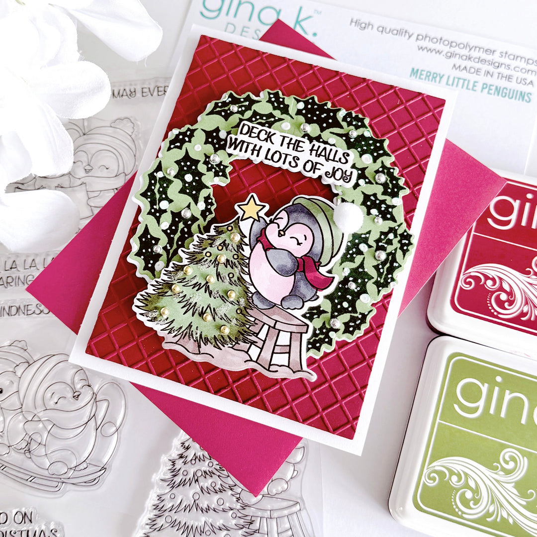A handmade Christmas card featuring a penguin sitting on a small stool beside a decorated tree, with the text 'Deck the Halls with Lots of Joy!'. The card is layered over a red textured background and pink envelope, with Gina K.