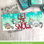A handmade Christmas card featuring penguins, snowflakes, and the text 'LET IT SNOW' in red, displayed on a light wood background with crafting tools like an ink pad and clear stamps.