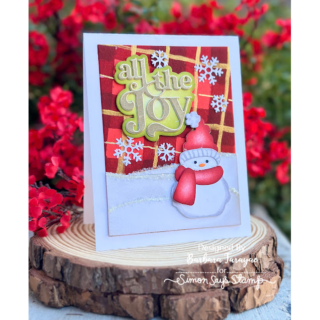 A handmade Christmas greeting card displayed on a rustic wood slice stand, with blurred red flowers in the background. The card features a white border, a red plaid pattern, the phrase 'all the joy' in green and gold glittery lettering, white snowflakes, and a 3D snowman wearing a red hat and scarf. Simon Says Stamp Bundled Snowman Wafer Dies 1527sd Barbara Tarayao | color-code:ALT04