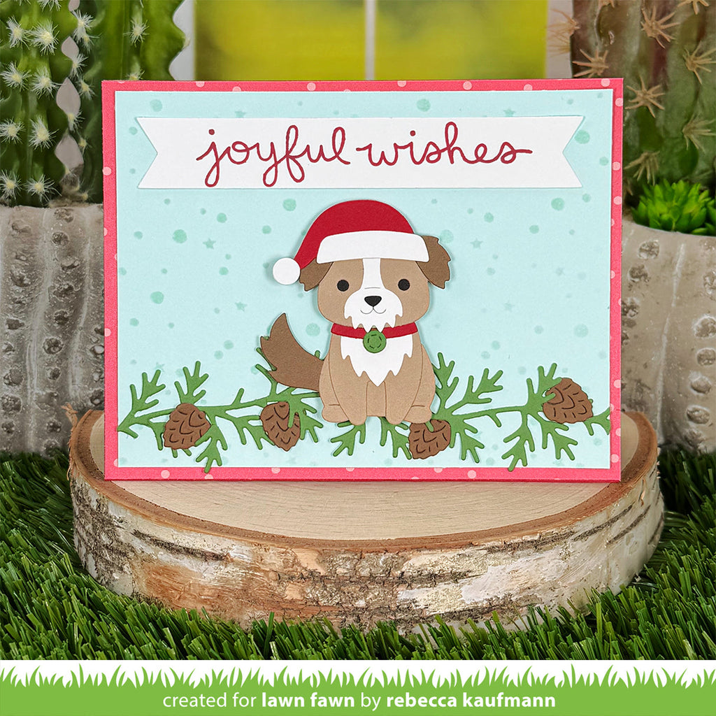 A handmade Christmas greeting card featuring a brown and white dog wearing a red Santa hat, set against a light blue background with green pine branches and pinecones. The card is displayed on a rustic wood slice surrounded by artificial grass and cacti.