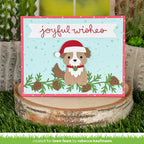 A handmade Christmas greeting card featuring a brown and white dog wearing a red Santa hat, set against a light blue background with green pine branches and pinecones. The card is displayed on a rustic wood slice surrounded by artificial grass and cacti.