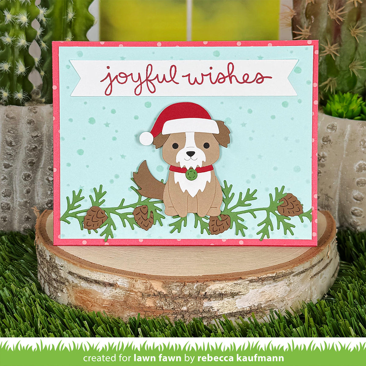 A handmade Christmas greeting card featuring a brown and white dog wearing a red Santa hat, set against a light blue background with green pine branches and pinecones. The card is displayed on a rustic wood slice surrounded by artificial grass and cacti.