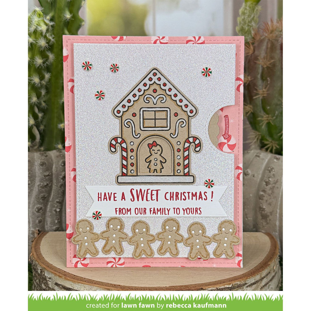 A handmade Christmas greeting card featuring a gingerbread house design, candy canes, and gingerbread men, displayed on a wooden slice with cacti in the background.