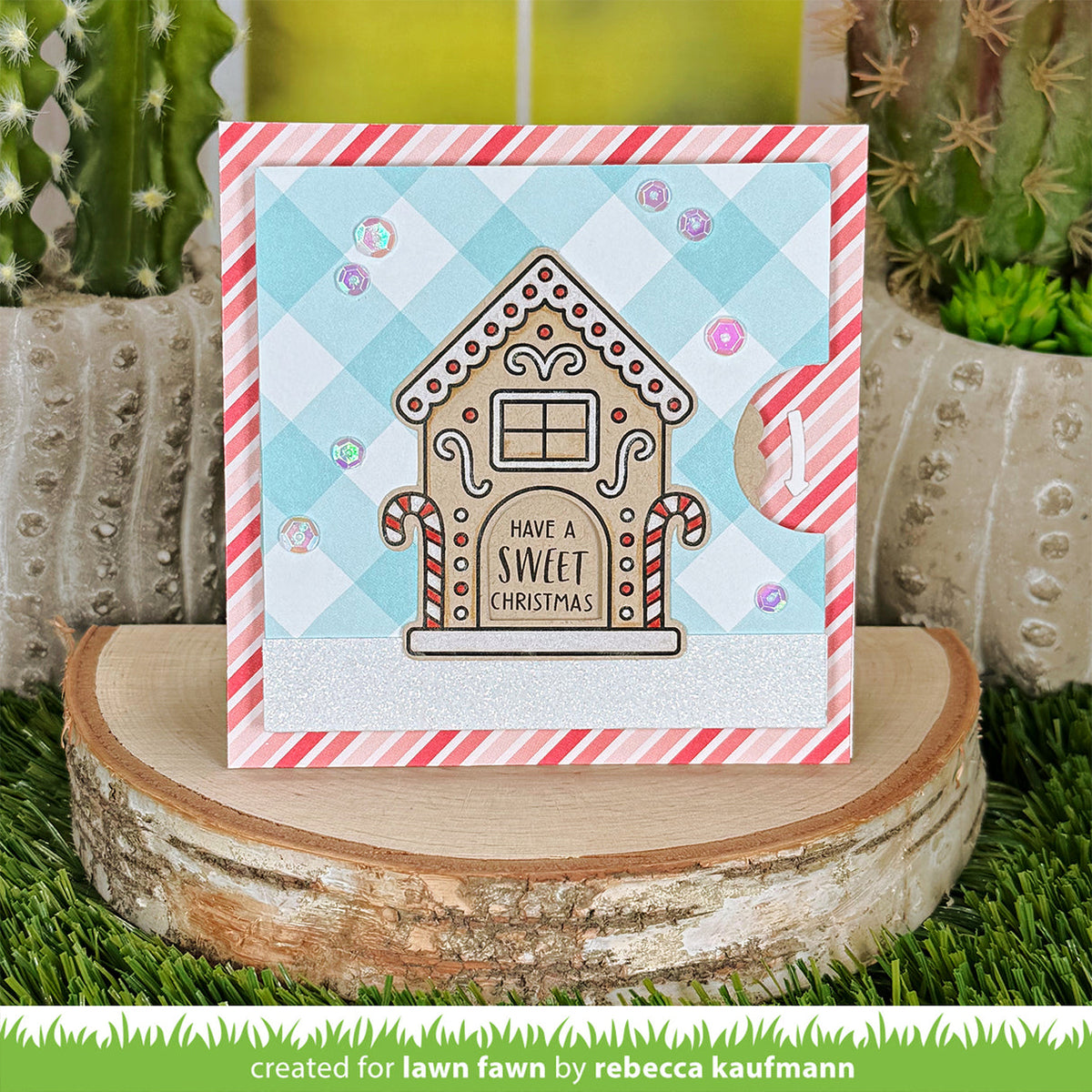 A handmade Christmas greeting card featuring a gingerbread house with the text 'HAVE A SWEET CHRISTMAS' on a light blue and white gingham background with red-and-white striped borders. The card is displayed on a rustic wood slice with cacti in the background.