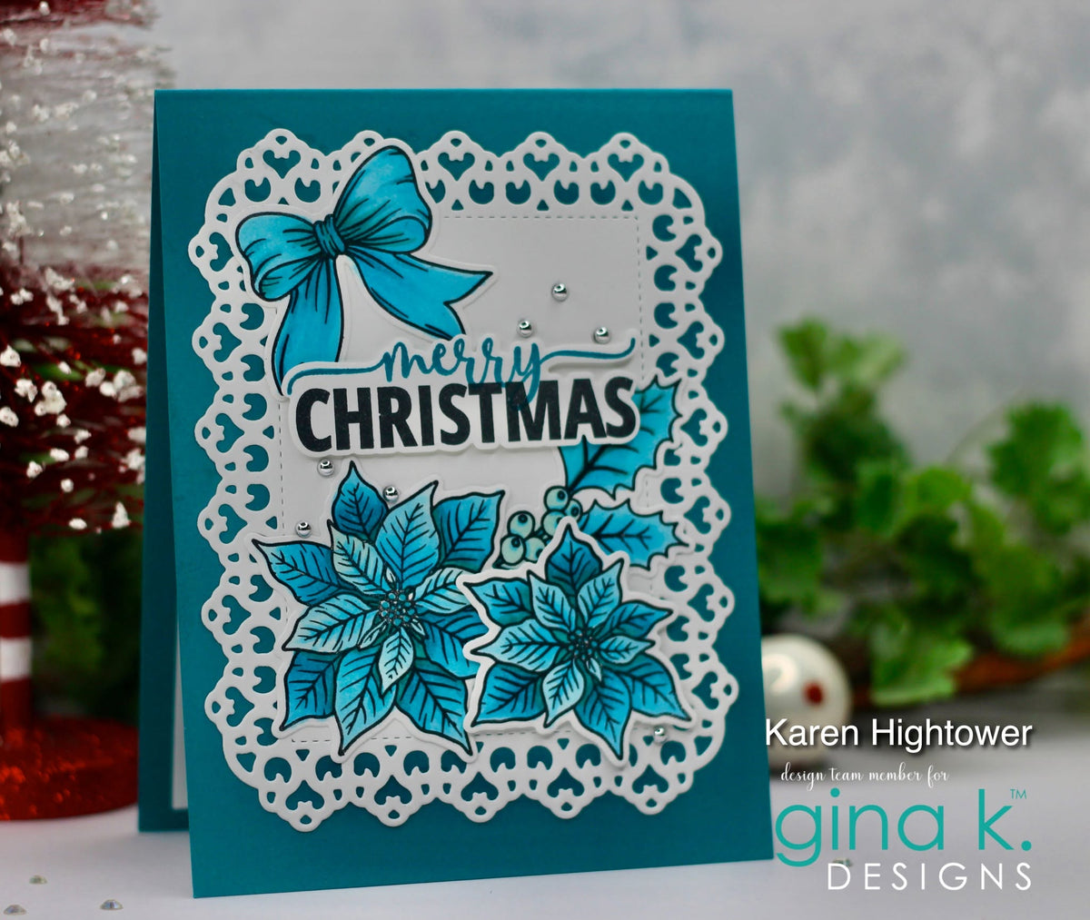 A handmade Christmas greeting card featuring a teal background, a white scalloped die-cut border, a turquoise bow, poinsettia flowers, and the words 'merry CHRISTMAS' in black script and block lettering. The card is photographed on a tabletop with blurred holiday decorations and greenery in the background.