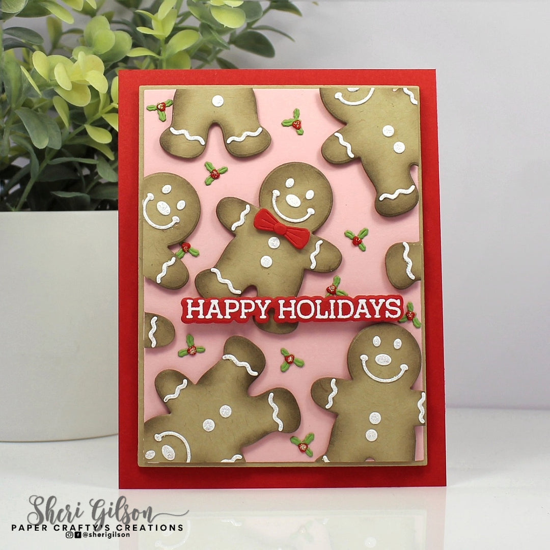 A handmade Christmas greeting card featuring gingerbread men cookies on a pink background with a red border and the words 'HAPPY HOLIDAYS' in white text on a red banner. The card is displayed on a white surface with a blurred green plant in the background.