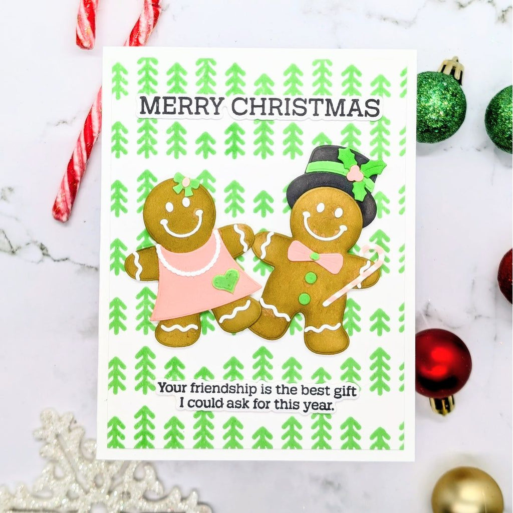 A handmade Christmas greeting card featuring two gingerbread cookies, one in a pink dress and one in a top hat with a green bow, on a white background with green pine tree patterns. The card is styled on a marble surface with holiday ornaments and candy canes.