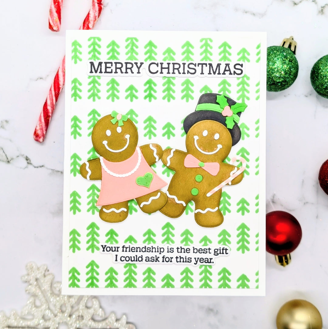 A handmade Christmas greeting card featuring two gingerbread cookies, one in a pink dress and one in a top hat with a green bow, on a white background with green pine tree patterns. The card is styled on a marble surface with holiday ornaments and candy canes.