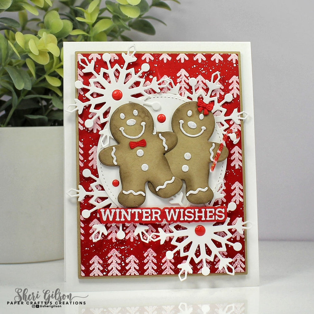 A handmade Christmas greeting card featuring two gingerbread men figures, white snowflakes, red patterned background, and the text 'WINTER WISHES' on a white cardstock, photographed with a plant in the background.