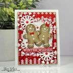 A handmade Christmas greeting card featuring two gingerbread men figures, white snowflakes, red patterned background, and the text 'WINTER WISHES' on a white cardstock, photographed with a plant in the background.