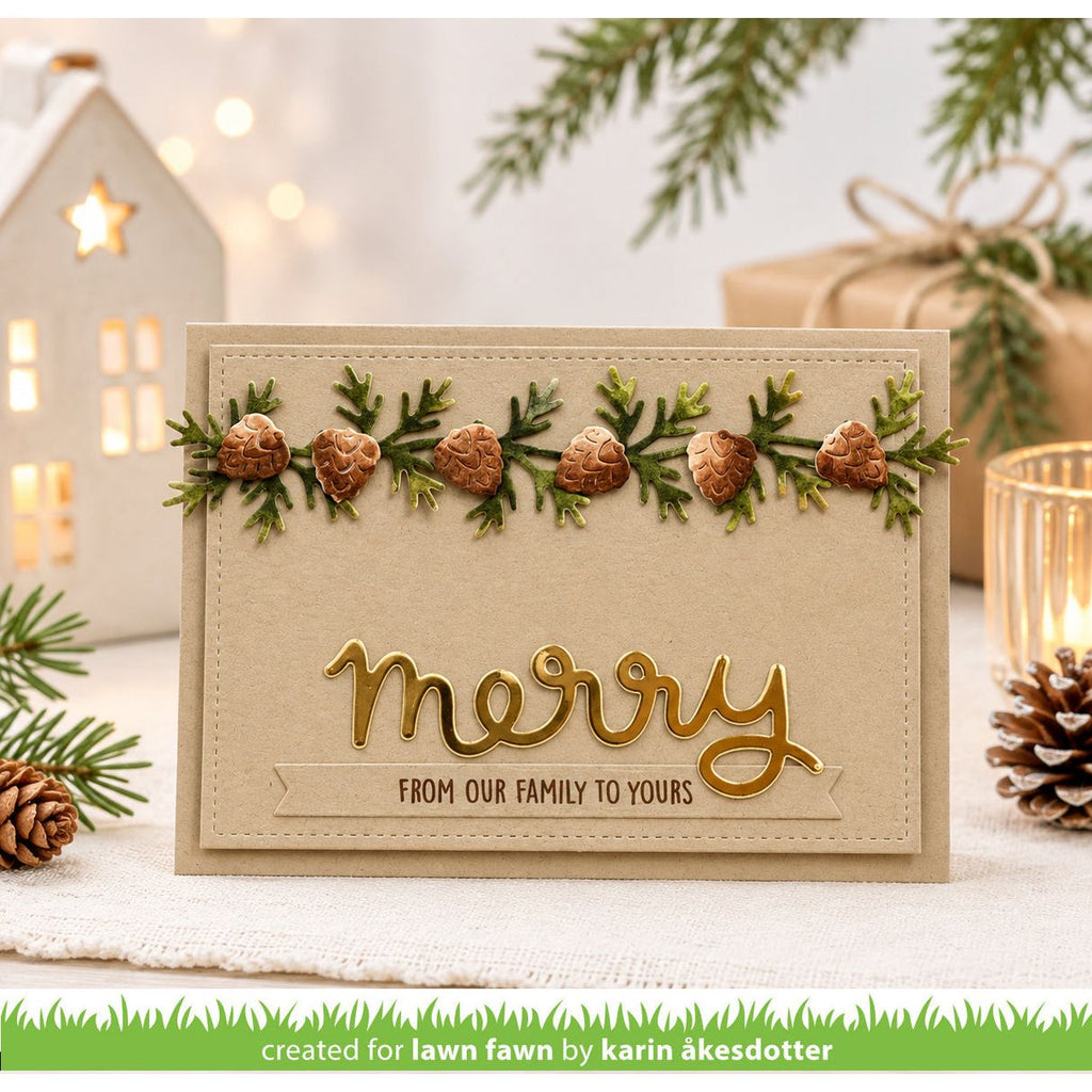 A handmade Christmas greeting card with a beige background, a green pine garland with pinecones across the top, and gold foil script reading 'merry' with the phrase 'FROM OUR FAMILY TO YOURS' below. The card is styled on a white surface with holiday decorations including pine branches, a lit candle, a small house ornament, and wrapped gift boxes in the background.