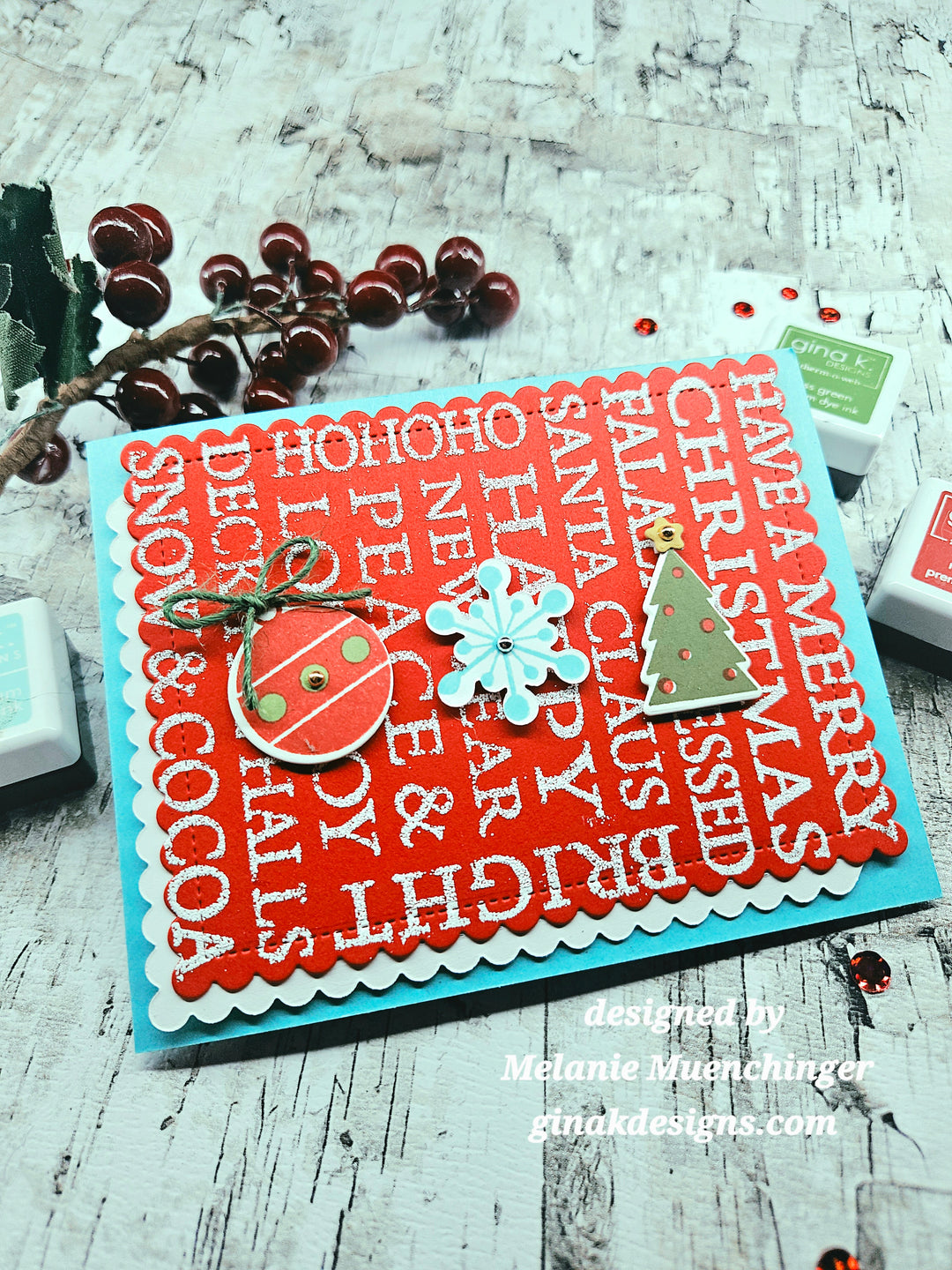A handmade Christmas greeting card with a bright red scalloped background printed with white holiday words like 'HOHOHO', 'SANTA', and 'CHRISTMAS', layered on a turquoise base. The card is decorated with a red ornament, a light blue snowflake, and a small green Christmas tree, styled on a rustic white wood surface with holiday berries and stamp ink pads.