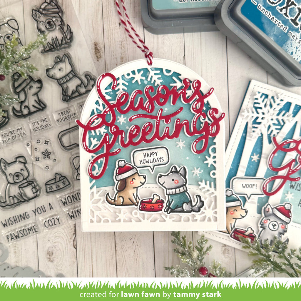 A handmade Christmas tag featuring layered die-cut paper with a winter dog illustration, red cursive 'Seasons Greetings' text, and a blue snowflake-patterned background. The card is styled on a whitewashed wood surface with clear stamped designs, ink pads, and additional card elements.