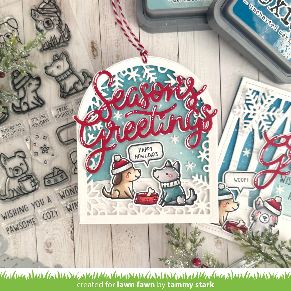 A handmade Christmas tag featuring layered die-cut paper with a winter dog illustration, red cursive 'Seasons Greetings' text, and a blue snowflake-patterned background. The card is styled on a whitewashed wood surface with clear stamped designs, ink pads, and additional card elements.