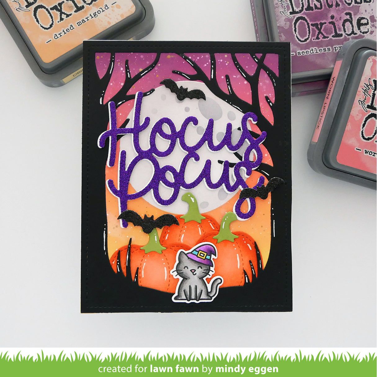 A handmade Halloween-themed card featuring layered black, purple, orange, and pink elements, including the words 'Hocus Pocus' in purple script over pumpkins, bats, a full moon, and a small cat sticker. Surrounding stamps are partially visible on a white surface. | color-code:alt1
