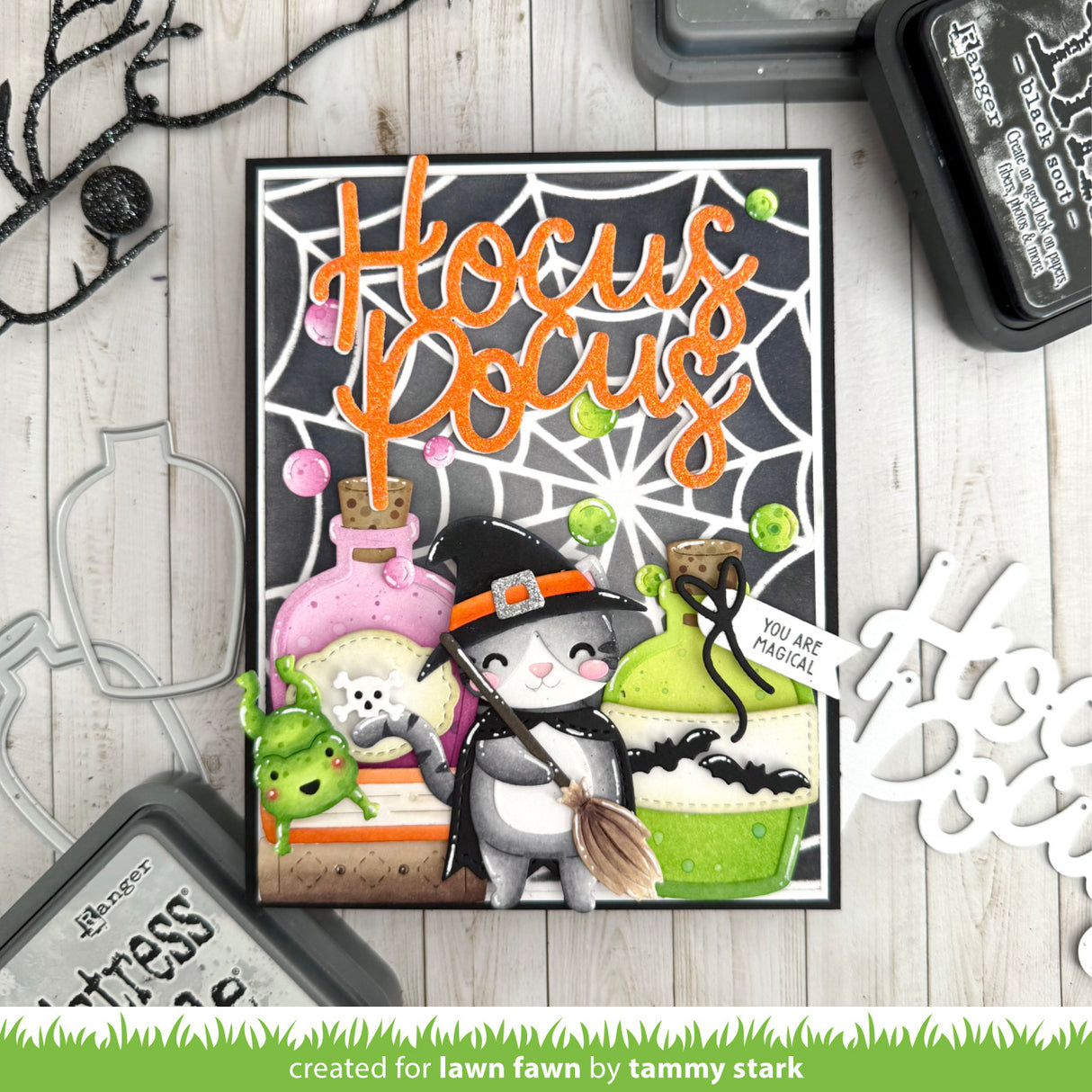 A handmade Halloween-themed card featuring the words 'Hocus Pocus' in orange script over a spiderweb background, with a witch cat, potion bottles, a broom, and small green monster figures. The card is photographed on a whitewashed wood surface with stamping tools and decorative elements around it.