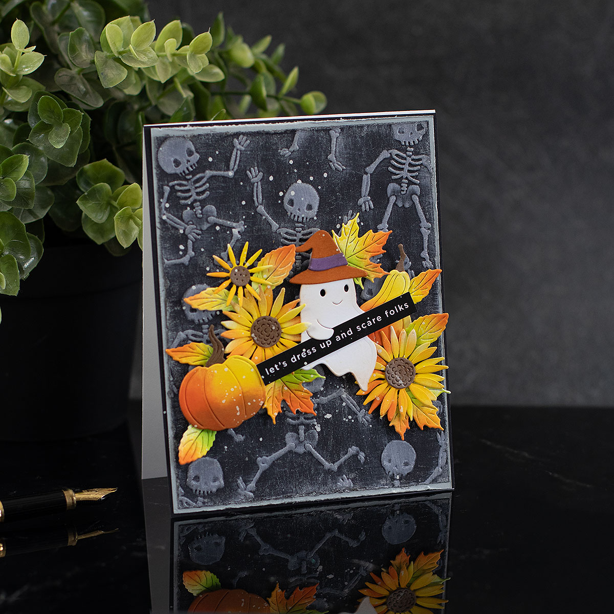 A handmade Halloween greeting card with a black and gray background, featuring layered paper skeletons, yellow sunflowers, a pumpkin, a ghost wearing a witch hat, and a banner that reads 'let's dress up and scare folks!'. The card is displayed standing upright on a dark reflective surface with a blurred plant in the background. Simon Says Stamp Embossing Folder Dancing Skeletons sf665 Spooky Ghost Halloween Card Bibi Cameron | color-code:ALT03