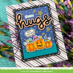 A handmade Halloween greeting card with layered black scalloped borders, a blue polka-dot background, orange glittery 'spooky hugs' lettering, and three orange pumpkins wearing purple witch hats. The card is displayed on a striped cardstock base with purple flowers around it. | color-code:alt2
