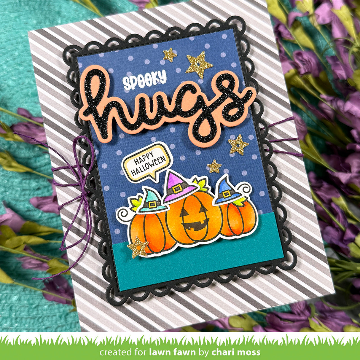 A handmade Halloween greeting card with layered black scalloped borders, a blue polka-dot background, orange glittery 'spooky hugs' lettering, and three orange pumpkins wearing purple witch hats. The card is displayed on a striped cardstock base with purple flowers around it. | color-code:alt2