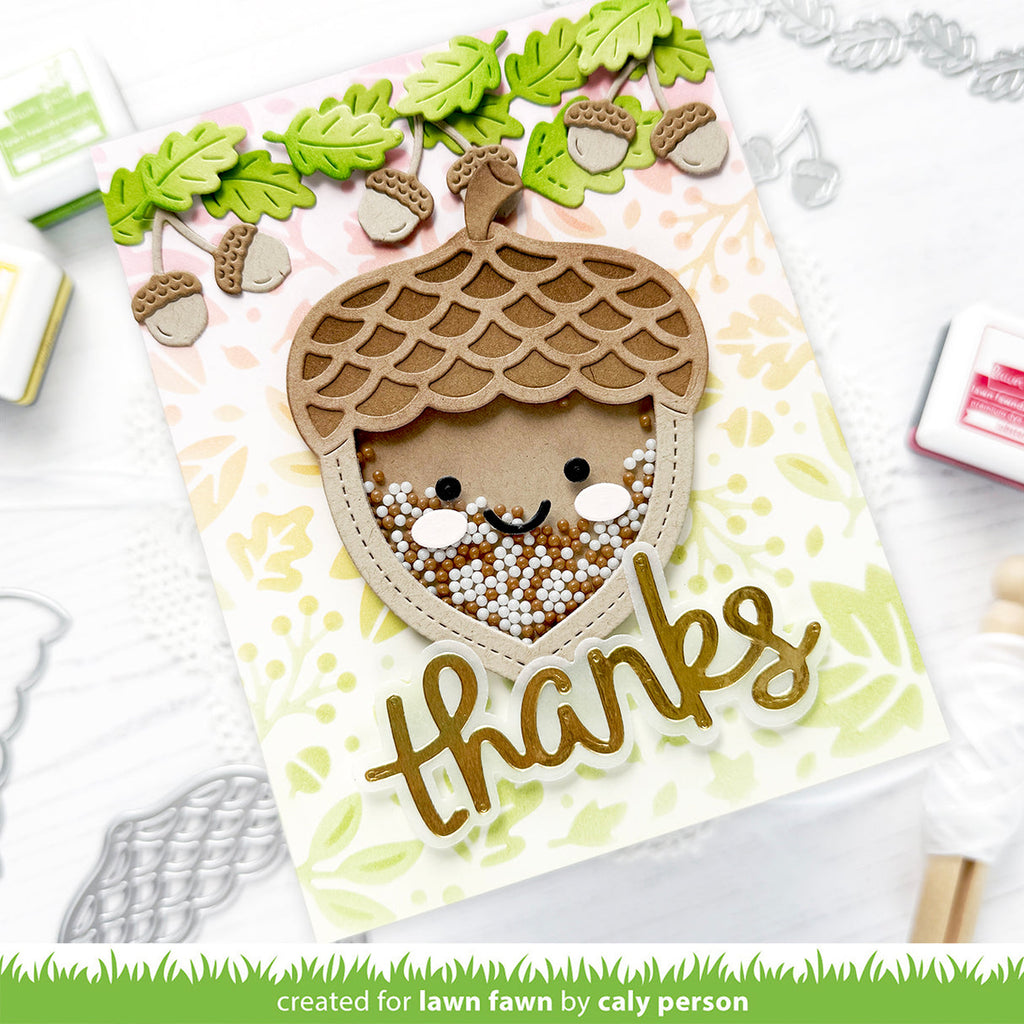 A handmade autumn-themed card featuring a cute acorn face with a smiling face peeking out from inside, decorated with green leaves and acorns along the top. The card has a gold 'thanks' script at the bottom and is displayed on a white background with crafting tools. | color-code:alt1