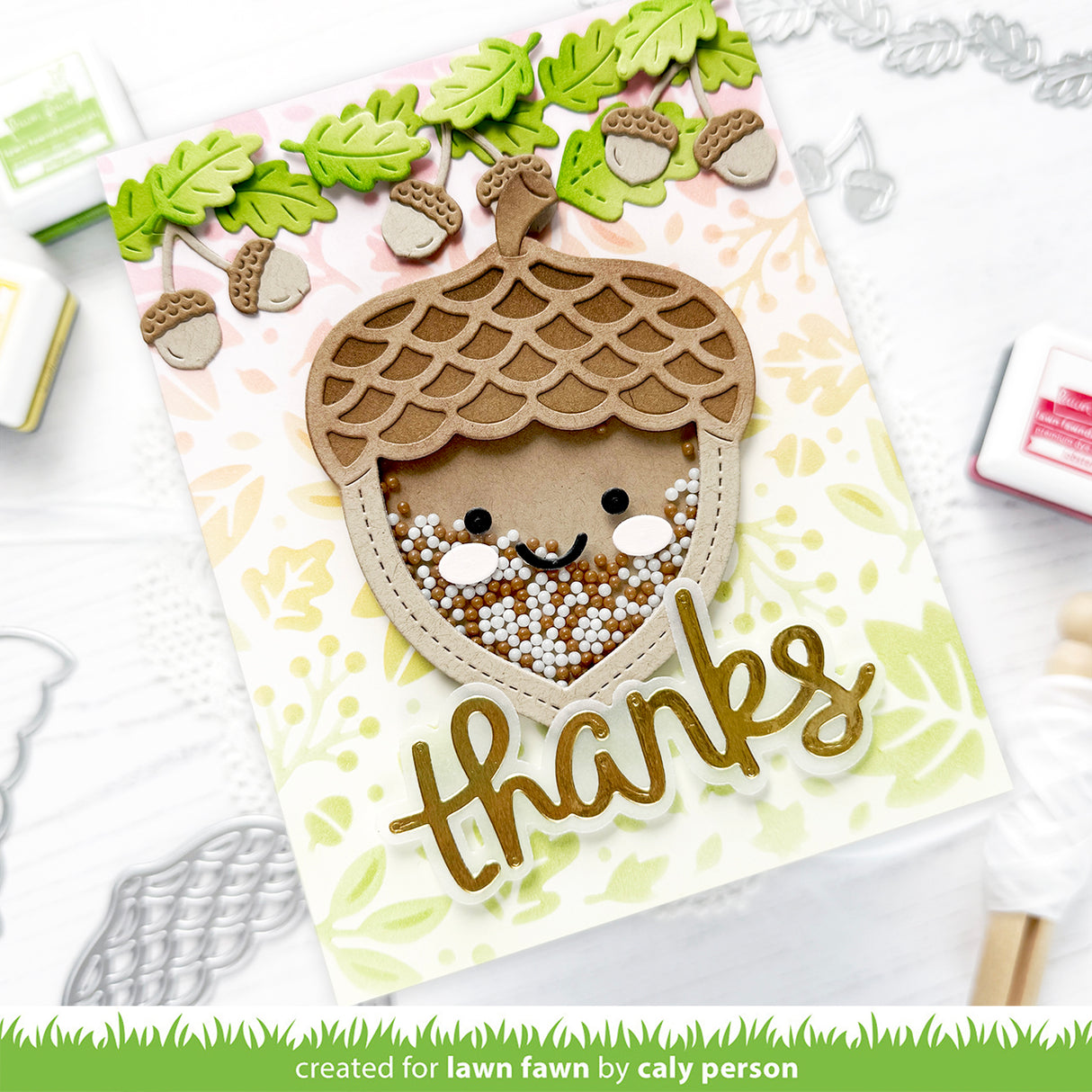A handmade autumn-themed card featuring a cute acorn face with a smiling face peeking out from inside, decorated with green leaves and acorns along the top. The card has a gold 'thanks' script at the bottom and is displayed on a white background with crafting tools. | color-code:alt1