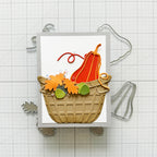 A handmade autumn-themed card featuring a tan basket filled with orange and yellow leaf-shaped embellishments, green acorns, and a red-orange pumpkin, displayed on a white grid background with metal craft dies.