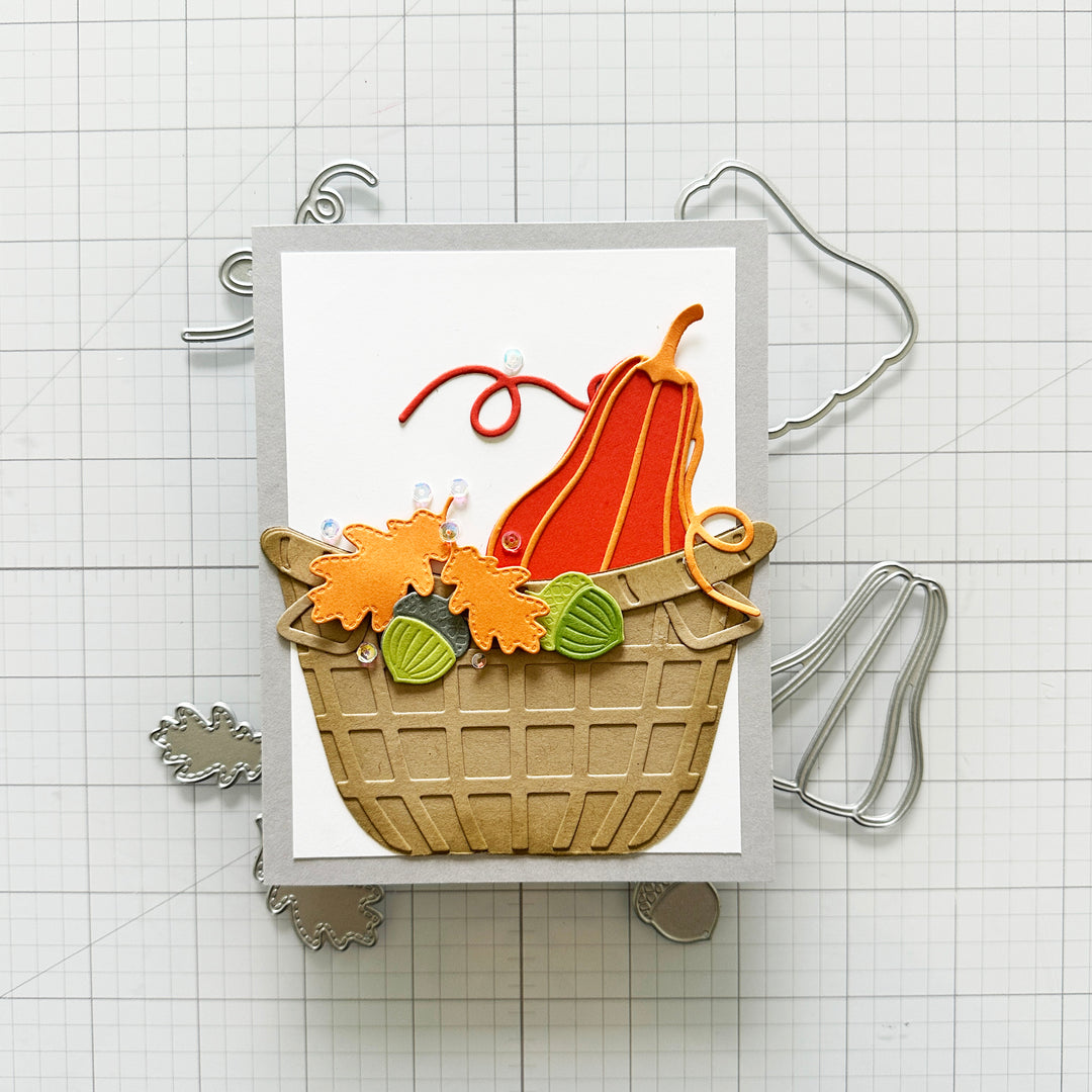 A handmade autumn-themed card featuring a tan basket filled with orange and yellow leaf-shaped embellishments, green acorns, and a red-orange pumpkin, displayed on a white grid background with metal craft dies.