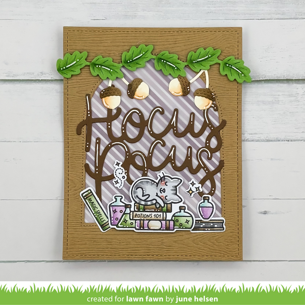 A handmade autumn-themed card with the words 'Hocus Focus' in brown script over a purple striped background, decorated with green leaves, acorns, a gray cat, potion bottles, and books. The card is photographed against a white wood plank background.