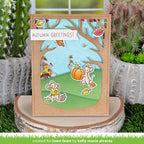 A handmade autumn-themed greeting card displayed on a wood slice stand, with cacti and artificial grass in the background. The card features layered die-cut designs including a light blue sky, green grass, brown tree branches, a pumpkin, two cartoon mice, and a white banner reading 'AUTUMN GREETINGS!'.