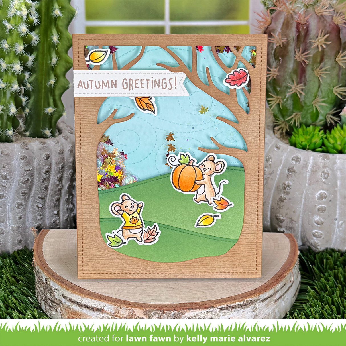 A handmade autumn-themed greeting card displayed on a wood slice stand, with cacti and artificial grass in the background. The card features layered die-cut designs including a light blue sky, green grass, brown tree branches, a pumpkin, two cartoon mice, and a white banner reading 'AUTUMN GREETINGS!'.