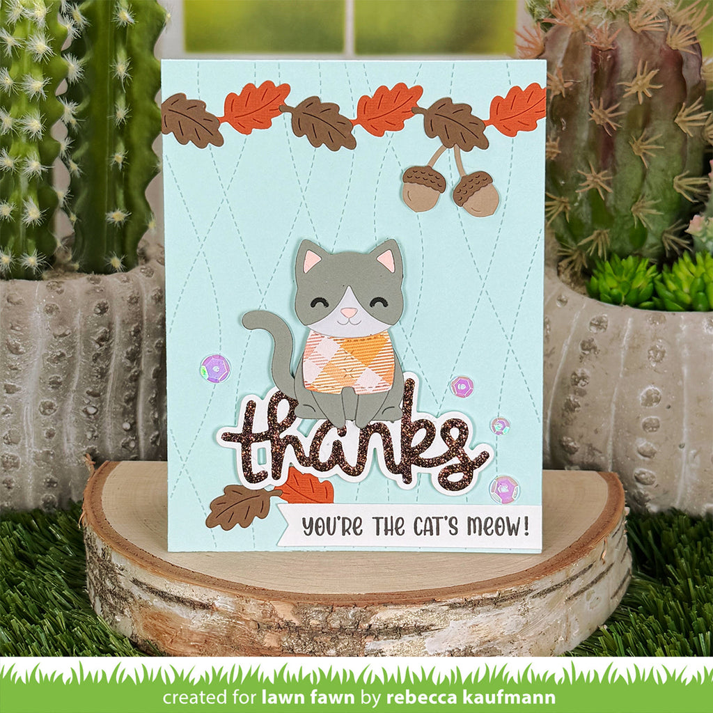 A handmade autumn-themed greeting card featuring a cute gray cat with an orange patterned sweater, acorns, and the words 'thanks' and 'you're the cat's meow!' displayed on a rustic wood slice stand, with cacti in the background.